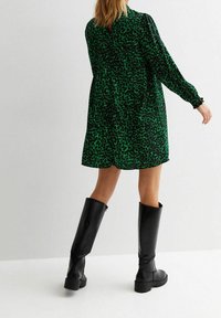 Zelené šaty s leopardím potiskem, dlouhými rukávy a uvolněným střihem, doplněné o černé kožené boty nad kolena s masivní podrážkou.