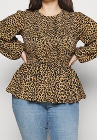 Frau in einem langärmligen Peplum-Top mit Leopardenmuster und gerafftem Oberteil, kombiniert mit hellblauen Jeans, die Hände in die Hüften gestützt.
