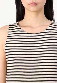Femme portant un haut blanc sans manches avec des rayures noires horizontales espacées uniformément, montrée des épaules aux lèvres sur un fond uni.