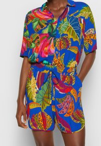 Donna che indossa una vivace tuta blu con motivi tropicali di foglie e fiori multicolori, bottoni davanti e una vita regolabile con coulisse.