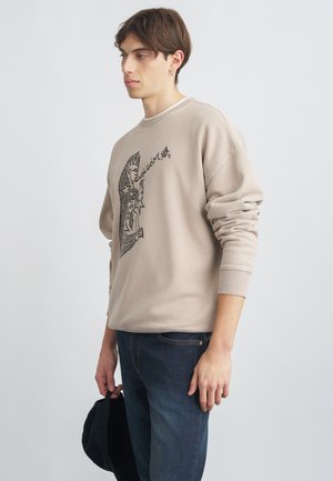 Sweatshirt beige à manches longues avec impression graphique noire sur le devant et le dos. Associé à un jean bleu foncé et tenant une casquette noire.