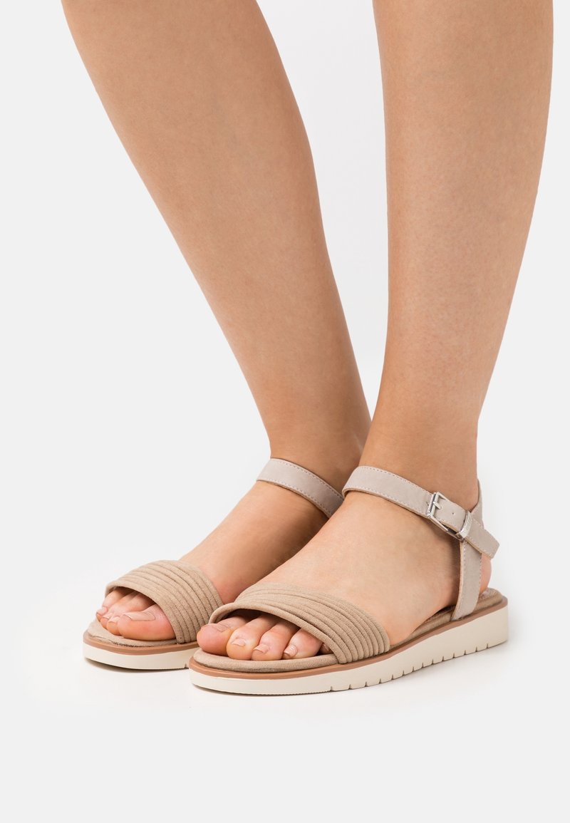 Beige-sandaalit, joissa on raidoitettu kangaspäällinen, säädettävä nilkkahihna ja pehmustettu pohja. Sisältää vaaleanvärisen teksturoidun lautasen.