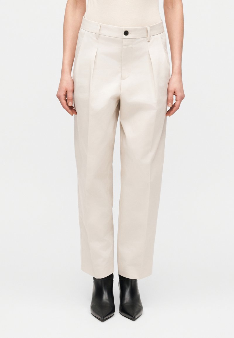 Beiget korkeavyötäröiset housut, joissa on laskokset, etutaskut ja napillinen kiinnitys. Valmistettu sileästä kankaasta, yhdistetty mustien nilkkureiden kanssa.