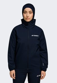 Námornícky modrá bunda Terrex s vysokým golierom, plným zipsom a kontrastným bielym logom. Vyrobená z hladkého, vodeodolného materiálu.