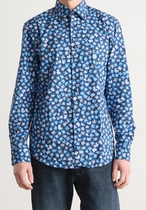 Homme portant une chemise ajustée à manches longues boutonnée avec un motif floral abstrait bleu, gris et violet clair et un jean sombre.