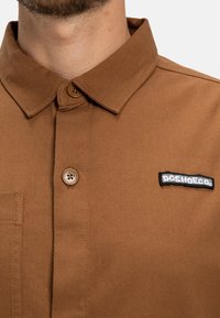Braun kariertes Button-Down-Hemd aus Baumwolle mit Kragen, einer Brusttasche und einem gestickten DC Shoes-Logo in Schwarz und Weiß auf der linken Brust.