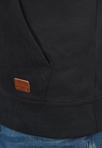 Black hoodie with a textured surface, featuring a leather logo patch and ribbed cuffs and hem. The pocket seam is visible on the left side.