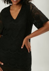 Schwarze Häkel-Tunika mit V-Ausschnitt und kurzen Ärmeln, die ein filigranes Muster und einen strukturierten Stoff zeigt, mit offenen Strickdetails.