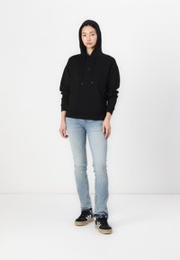Sweatshirt à capuche noir avec poche avant et cordons de serrage, associé à un jean slim-fit bleu clair et des baskets noires avec des rayures blanches.