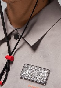 Grijze button-up overhemd met een gestructureerde stof, een kraag en een zwarte naambord met logo. Rode accenten en trekkers zijn zichtbaar.