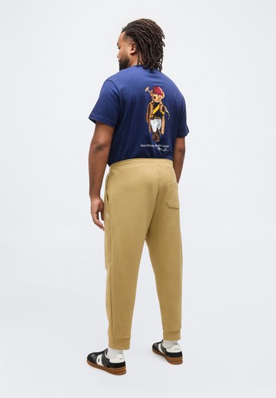 Άνδρας με κότσους ντρέντλοκ, φορώντας μπλε ναυτικό T-shirt με γραφικό αρκούδας πόλο, μπεζ φόρμα, λευκές κάλτσες και ασπρόμαυρα αθλητικά παπούτσια, όρθιος σε λευκό φόντο.