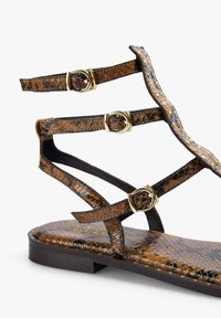 Sandalias marrones con patrón de serpiente y suelas planas, que presentan tres correas ajustables y hebillas en tono dorado. Parte superior de textura suave.