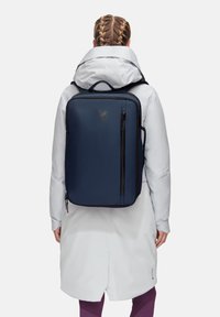 Sac à dos navy de forme rectangulaire, avec une poche avant et des accents de zip noir, porté sur une veste gris clair et une couche extérieure blanche.