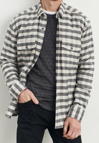 Button-up skjorte med grå og hvide vandrette striber, lavet af flannel. Har to forlommer og en krave. Bæres over en mørk sweater.