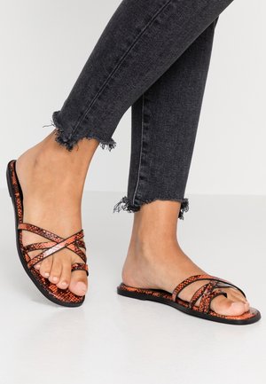 Pies con sandalias planas de estampado de serpiente en naranja y negro con tiras delgadas, combinadas con jeans ajustados negros con dobladillo deshilachado.