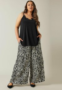 Zwarte mouwloze top met kantdetail, gecombineerd met losse, wijde broek met een wit bloemenpatroon op een zwarte achtergrond.
