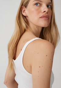 Top bianco senza maniche indossato da una donna, con spalle e capelli in mostra. Tessuto liscio, design aderente, con macchie di pelle visibili su spalle e schiena.