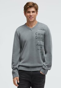 Grauer Strickpullover mit V-Ausschnitt, gerippten Bündchen und Saum. Verziert mit schwarzem und dunkelgrauem Aufdruck. Wird über schwarzen Hosen getragen.