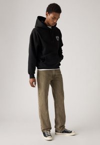 Haut à capuche noir avec poche avant, cordon de serrage réglable et logo sur la poitrine ; associé à un pantalon beige à jambes droites et des baskets noires.
