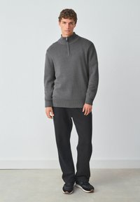 Donkergrijze gebreide trui met een hoge kraag en een halve rits, gecombineerd met zwarte joggingbroek en zwarte sneakers. Gladde textuur, casual pasvorm.