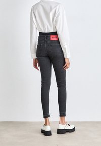 Persona que lleva pantalones ajustados negros de cintura alta con la etiqueta roja "HUGO", una camisa blanca de manga larga y deportivas blancas con plataforma, de pie sobre un suelo claro.