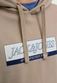 Tanowy hoodie z grafiką z przodu zawierającą tekst "JACK&JONES" i "SIN NINETEEN NINETY". Sznurki i miękka tekstura.