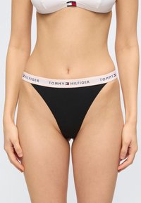 Femme portant une culotte noire taille haute avec une ceinture élastique blanche brandée "Tommy Hilfiger" et un soutien-gorge blanc avec logo.
