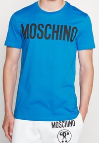 Osoba v modré košili s krátkým rukávem, na které jsou černá písmena „MOSCHINO“, a v bílých kalhotách s brandingem a logem „MOSCHINO“ na levé kapse.