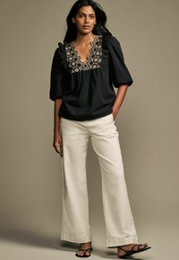 Blusa nera con scollatura a volant e ricami beige intricati, abbinata a pantaloni bianchi a gamba larga e sandali neri con cinturino.