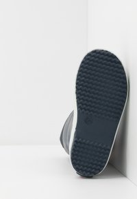 Tmavá gumová podrážka boty s texturovaným, cik-cak vzorem a bílým akcentem kolem okraje. Lehce zakřivený tvar, žádné viditelné logo nebo vzory.