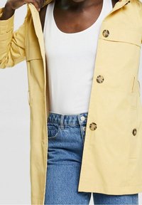 Imperméable jaune clair avec capuche, doté de deux grands boutons, poches latérales et coupe décontractée, associé à un débardeur blanc et un jean bleu.