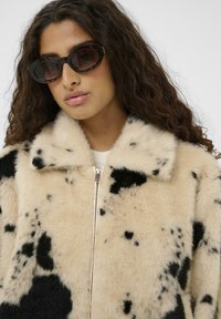 Umetno krzno s kremno osnovo, okrašeno s črnimi pikastimi vzorci, z zadrgo in širokim ovratnikom. Model nosi ovalna črna sončna očala.