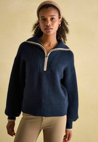 Dunkelblauer Pullover mit Halbreißverschluss-Kragen, geripptem Saum und weiten Ärmeln. Verfügt über einen kontrastierenden beigen Reißverschluss und einen strukturierten Stoff.