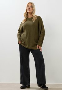 Oliwkowy sweter z dzianiny o luźnym kroju, okrągłym dekolcie i guzikami po bokach. Połączony z szerokimi czarnymi spodniami i czarnymi butami wsuwanymi.