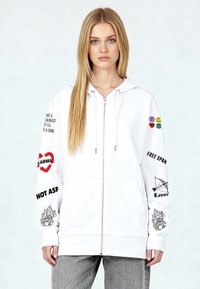 Felpa con cappuccio bianca con zip frontale, tasche anteriori e stampe grafiche. Presenta testi ed emoji colorate sulle maniche e un disegno a forma di cuore.