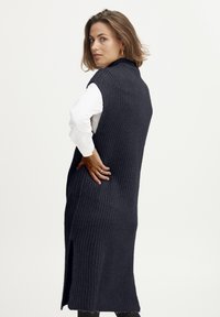 Langer, ärmelloser, dunkelblauer Strick-Cardigan mit strukturiertem Muster, seitlichen Schlitzern und hohem Kragen. Gestylt über einem weißen Hemd.