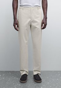 Chinos beige con un design a gamba dritta, realizzati in tessuto liscio. Presentano una chiusura con bottone e sottili tasche laterali. Abbinati a scarpe scure.