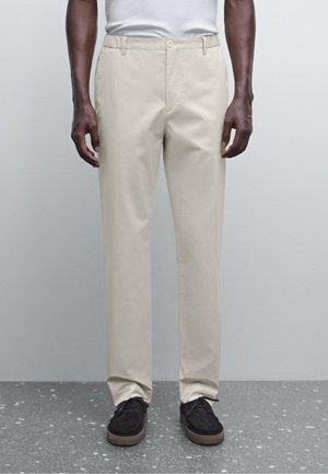 Beige chinos with a straight-leg design, made from smooth fabric. Features a button closure and subtle side pockets. Paired with dark shoes.