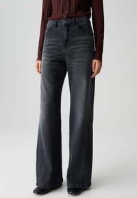 Personne portant un jean large en denim noir délavé �à taille haute, associé à des chaussures noires et un haut long à manches ribbées bordeaux.