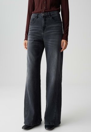 Persoon met hoge taille, wijd uitlopende, zwarte gewassen denim jeans, gecombineerd met zwarte schoenen en een wijnrode geribbelde lange mouwen top.