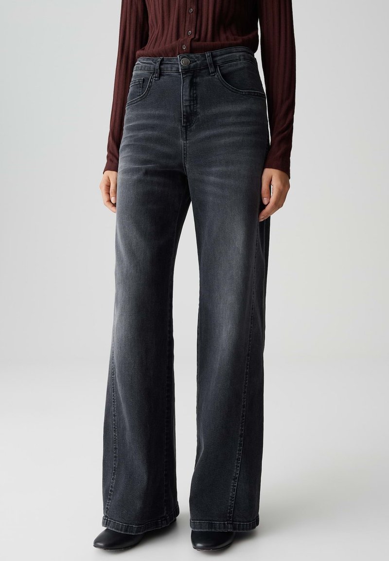 Personne portant un jean large en denim noir délavé à taille haute, associé à des chaussures noires et un haut long à manches ribbées bordeaux.