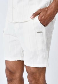 Shorts bianchi lavorati a maglia con un motivo testurizzato, tasca laterale e un accento metallico vicino alla tasca. Il tessuto è morbido e traspirante.