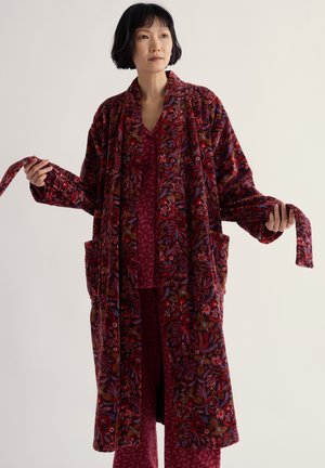 Femme tenant une large ceinture portant une longue robe fleurie rouge foncé à motifs sur un pyjama rouge assorti à motifs.