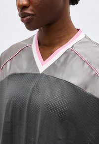 Κοντινή λήψη ενός ατόμου που φοράει γκρι αθλητική φανέλα με ροζ και λευκή γαζωτή V-λαιμόκοψη και ροζ ραφή στους ώμους.