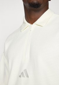 Bela pulover s četrtno zadrgo iz teksturirane tkanine, ki ima ovratnik in sivi adidas logo na prsih. Preprost, minimalističen dizajn.