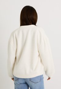 Sudadera de felpa blanca con un corte holgado, que presenta una superficie ligeramente texturizada y puños acanalados, combinada con jeans azul claro.
