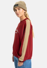 Rode sweatshirt met beige zijkanten, geribbelde manchetten en kraag. Gecombineerd met een beige geribbelde beanie. Model staat tegen een effen achtergrond.