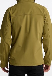 Man som bär en mjuk skaljacka i olivgrön färg med långa ärmar, synliga sömmar och huva, sedd bakifrån mot en vit bakgrund.