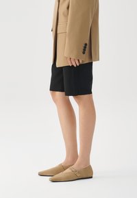 Béžové nubukové slip-on boty s špičatou špičkou a malým sponkovým detailem. Nosit s černými šortkami a béžovým oversized sako.
