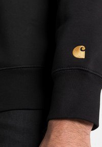 Felpa nera con polsini e orlo a costine, presenta un logo ricamato in oro sulla manica sinistra. Il materiale sembra morbido e leggermente testurizzato.